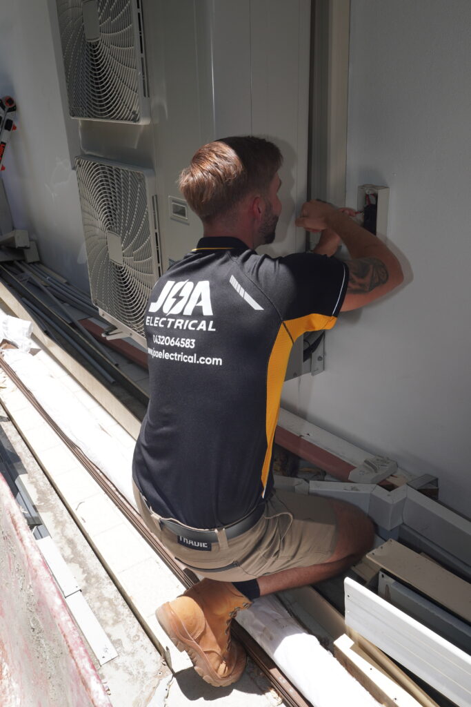 Licensed electrician installing outdoor electrical components beside air conditioning units